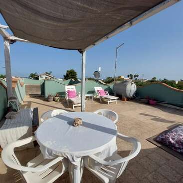 Das Bild zeigt eine ruhige Dachterrasse mit einem weißen Tisch und Stühlen, umgeben von einer hellblauen Wand und einem klaren blauen Himmel im Hintergrund an einem sonnigen Tag.