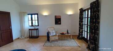 Este acogedor salón cuenta con un sillón de mimbre, una mesita con fruta y vino, dos ventanas con cortinas de flores, iluminación suave y suelo de baldosas.