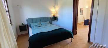 Un acogedor dormitorio cuenta con una cama gris con ropa de cama azul, mesillas de noche, lámparas de pared, suelo de baldosas, y conecta con una luminosa habitación contigua con decoración moderna.