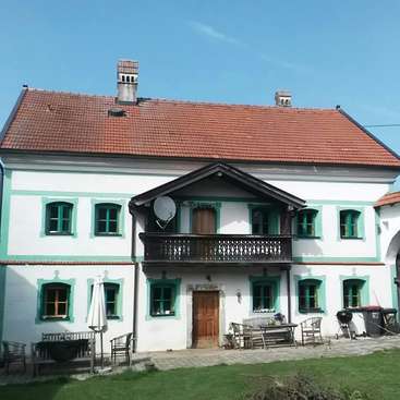 The image depicts a two-story white house with green trim and a red roof, featuring a balcony and patio area, set against a clear blue sky.