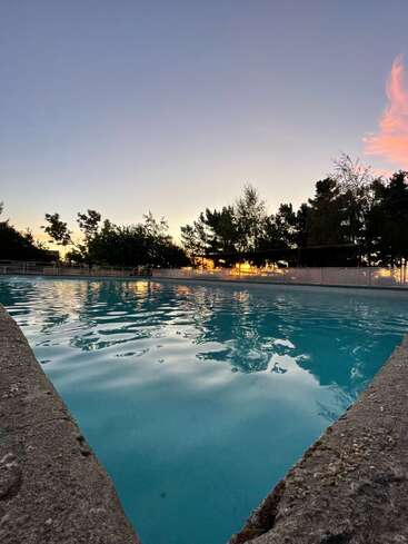 Ein ruhiger Swimmingpool spiegelt die sanften Farben eines Sonnenuntergangs wider. Bäume umgeben den Pool, und der Himmel wechselt von Blau zu warmen Abendtönen. Friedliche Szene.