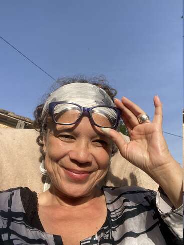 Une femme souriante, baignée par la lumière du soleil, vêtue d'une chemise à motifs, d'un foulard argenté et de lunettes posées sur le front, profite d'un moment de détente sous un ciel bleu limpide.