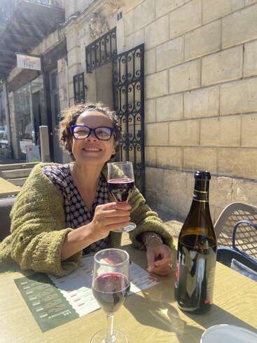 Une femme à lunettes sourit, un verre de vin rouge à la main, à la terrasse d'un café. Une bouteille et un autre verre sont posés sur la table, baignée par la lumière du soleil.
