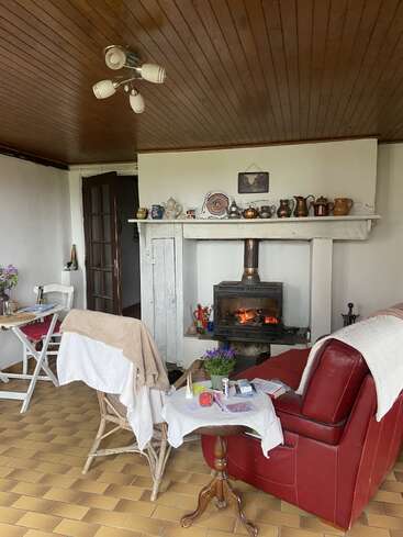 Un salon chaleureux avec un poêle à bois, un fauteuil rouge, des petites tables, une décoration rustique, des poteries sur la cheminée, un sol carrelé et un plafond en bois, qui respire la chaleur.