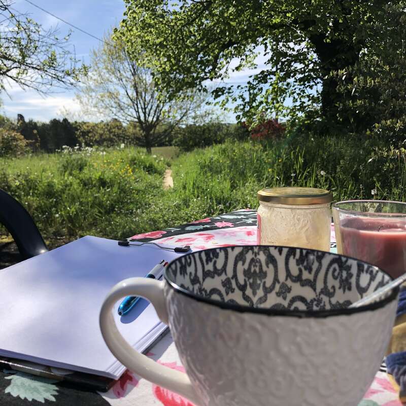 La imagen representa un entorno sereno al aire libre, con una mesa con un mantel de flores, un cuaderno, una taza y velas, en un pintoresco campo de hierba y árboles.