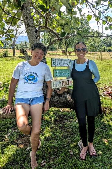 Zwei Frauen stehen lächelnd im Freien an einem Baum. Die eine lehnt sich an einen Ast, die andere hält ein Schild mit der Aufschrift "Willkommen an unserem Happy Place". Sonnige, friedliche Umgebung.