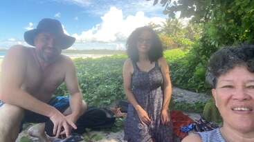 Drei Menschen entspannen sich an einem Strand, umgeben von üppigem Grün. Sie lächeln und freuen sich über das sonnige Wetter. Der Himmel ist blau mit flauschigen Wolken.