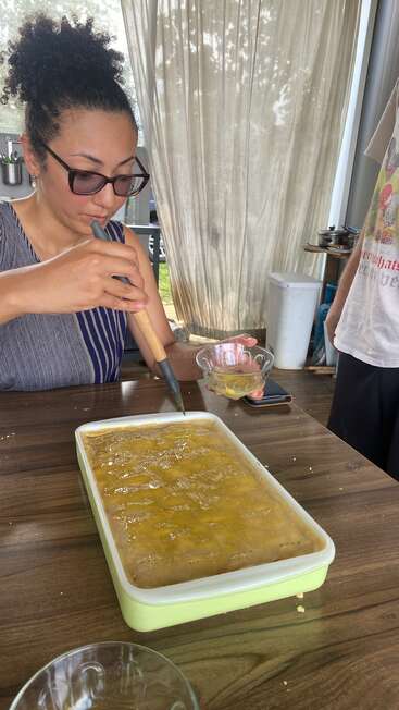 Eine Frau mit Sonnenbrille schleudert mit einem Metallwerkzeug Honig aus einer Wabenschale und sammelt ihn in einer kleinen Glasschale auf einem Holztisch.