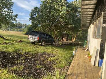Ein schwarzer Geländewagen parkt auf einem grasbewachsenen Hof in der Nähe einer Holzveranda. Große Bäume spenden Schatten, und weiter hinten steht ein kleines Gebäude. Eine friedliche, ländliche Umgebung.