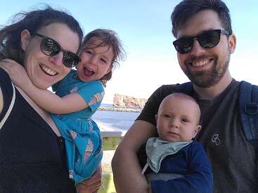 Das Bild zeigt eine vierköpfige Familie, die an einem sonnigen Tag für ein Selfie posiert, mit einem malerischen Gewässer und einer felsigen Küstenlinie im Hintergrund.