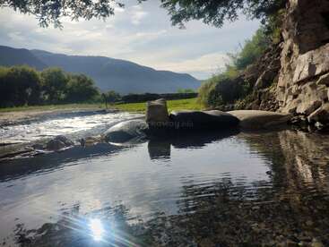 A peaceful scene featuring a clear stream with stones, surrounded by lush greenery, trees, and distant mountains under a bright sky with soft sunlight reflecting.