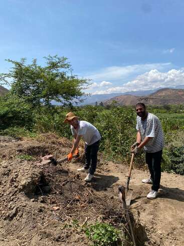 Two men are digging with shovels in a rural area surrounded by greenery and mountains under a blue sky with scattered clouds, working together outdoors.