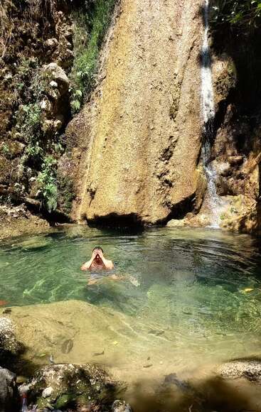 A person is swimming in a clear, shallow natural pool at the base of a small waterfall, surrounded by rocks, greenery, and sunlight. Peaceful nature scene.