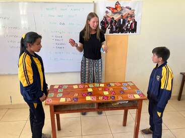 Un enseignant se tient entre deux élèves près d'une table recouverte de cartes colorées. Ils semblent jouer à un jeu éducatif dans une salle de classe.