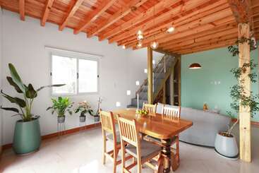 Uma sala de estar e jantar moderna e bem iluminada com teto de madeira, plantas de interior, móveis de madeira, paredes em tons pastéis, uma escada e iluminação exclusiva, criando uma atmosfera aconchegante.