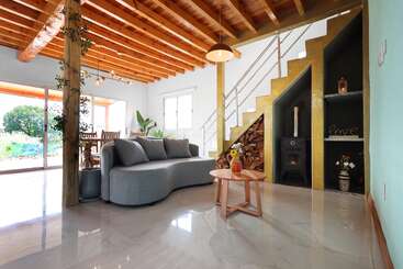 Esta sala de estar aconchegante apresenta um sofá cinza moderno, vigas de madeira no teto, luz natural abundante, um fogão a lenha, armazenamento inteligente embaixo da escada, plantas e uma decoração elegante.