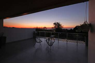 Um terraço tranquilo ao pôr do sol tem duas cadeiras modernas e uma mesa, com vista para as árvores e a paisagem aberta, com cores quentes iluminando o céu noturno.
