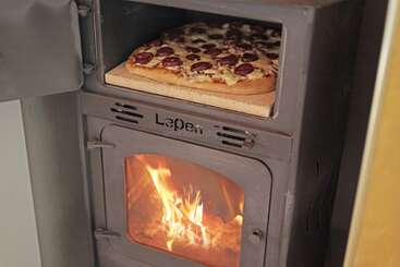 Uma pizza de aparência deliciosa coberta com queijo e linguiça está sendo assada em uma pedra dentro de um forno a lenha, com um fogo visível queimando abaixo.