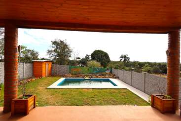 Um pátio coberto tem vista para um quintal cercado com uma piscina quadrada, gramado, galpão de madeira, vasos de plantas e árvores ao redor da área. O ambiente é tranquilo.