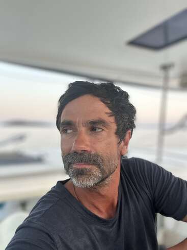 A man with short dark hair and a beard gazes to the side while sitting on a boat. The background is blurred, suggesting water and sky.