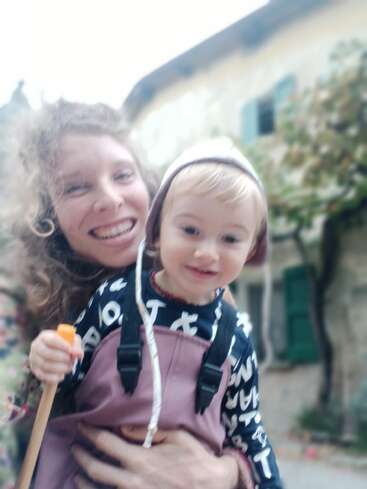 A happy woman and a small child smile at the camera outdoors. The background is blurred, showing a building with green windows and some greenery.