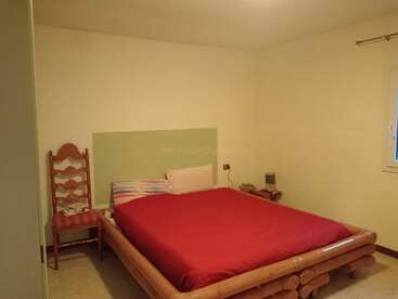 This simple bedroom features a wooden bed with a red blanket, a single chair, nightstand with speaker, minimal wall decorations, and a window for natural light.