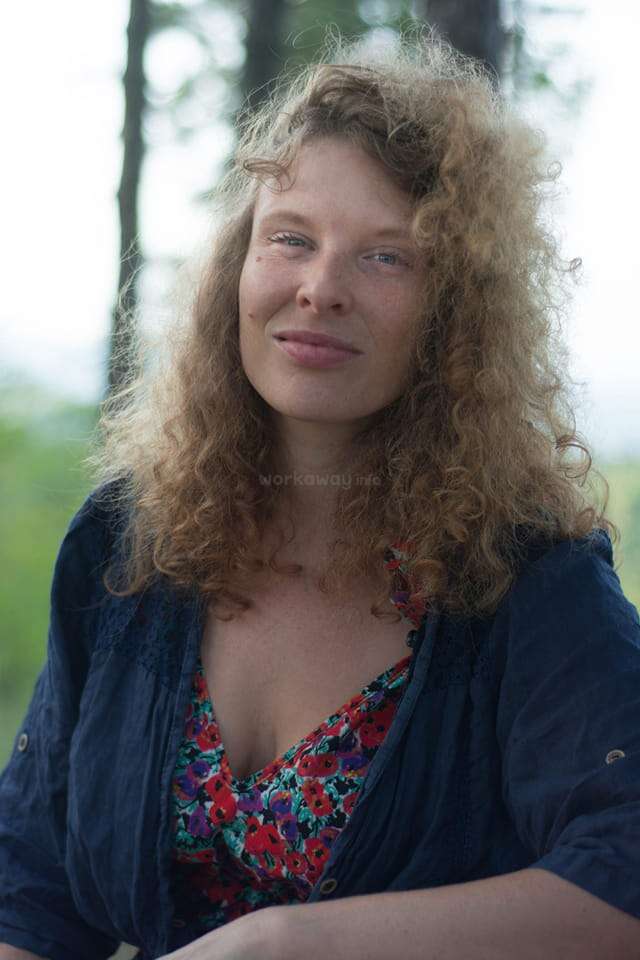A woman with curly, light-brown hair smiles softly at the camera. She is outdoors with a blurred, green and forested background, exuding natural beauty and calmness.