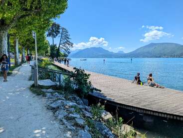 Die Menschen spazieren auf einem von Bäumen gesäumten Weg an einem ruhigen See entlang. Andere entspannen sich auf einem Holzsteg und genießen die Sonne und die atemberaubenden Berge im Hintergrund.