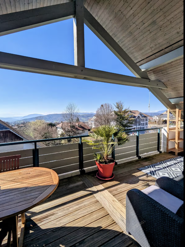 Ein gemütlicher Holzbalkon bietet einen herrlichen Blick auf die Berge und Dächer. Hier gibt es Sitzgelegenheiten im Freien, eine getopfte Palme, einen gemusterten Teppich und einen runden Holztisch.
