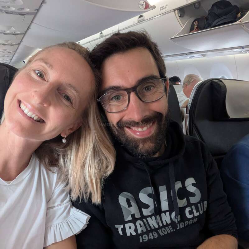 Ein glückliches Paar sitzt eng zusammen in einem Flugzeug und lächelt in die Kamera. Die Frau trägt ein weißes Hemd, der Mann eine Brille und einen Kapuzenpulli.