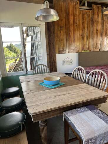 Un comedor rústico con una mesa de madera, sillas desparejadas, una lámpara de metal, cojines verdes, una ventana con vistas al campo y un centro de mesa decorativo con un cuenco azul.