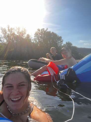 Three friends are floating on inner tubes in a river, smiling and enjoying the sunshine. The atmosphere is relaxed with trees and blue sky in the background.