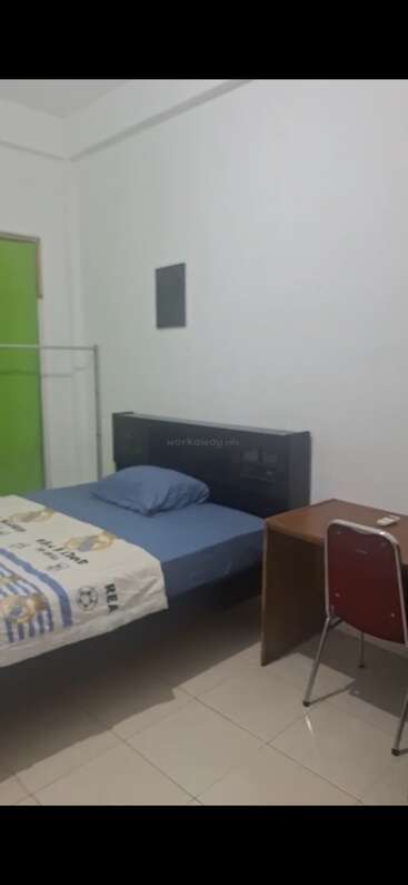 This image shows a simple bedroom with a single bed, blue bedding, a Real Madrid blanket, wooden desk, red chair, green door, and a clothing rack.