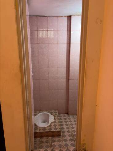 Esta imagen muestra un cuarto de baño pequeño con paredes alicatadas en rosa y suelo estampado, con un inodoro blanco de estilo turco. La iluminación parece tenue y las paredes necesitan limpieza.