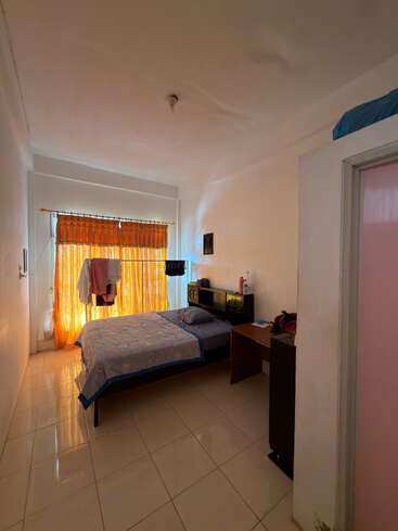 Una habitación pequeña y sencilla con una cama individual, cortinas naranjas, un escritorio, suelo de baldosas y una decoración minimalista. La luz del sol ilumina suavemente el espacio a través de la ventana.
