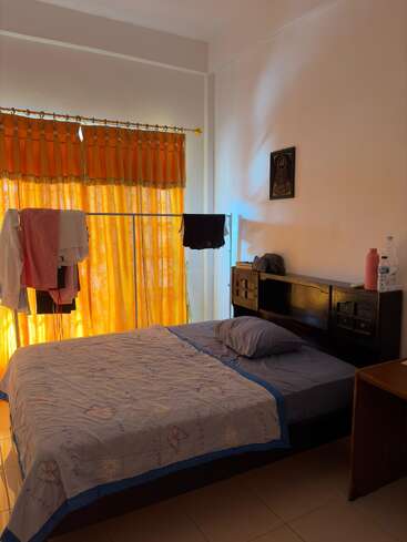 Una habitación sencilla con una cama bien hecha, ropa de cama morada, cortinas naranjas que filtran la luz del sol, ropa tendida en un tendedero y un cabecero de madera con algunos objetos encima.