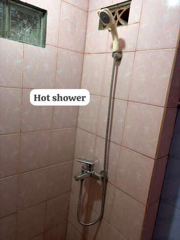 Esta imagen muestra una ducha de agua caliente en un cuarto de baño con azulejos rosas, un cabezal de ducha de mano, una pequeña ventana y el texto «Ducha de agua caliente» encima.