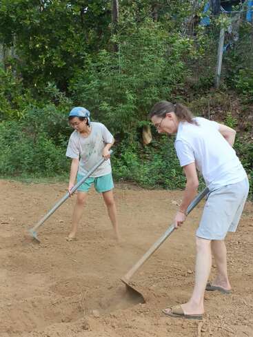 Dos personas trabajan juntas al aire libre, utilizando azadas para labrar o nivelar la tierra. Parecen concentradas, rodeadas de plantas verdes y vestidas con ropa cómoda e informal.