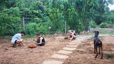 Three people are squatting and working on the ground, possibly gardening, with bowls beside them. A dog stands on a stone path, surrounded by lush greenery.