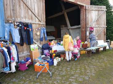 A small outdoor flea market with clothes, toys, and household items displayed outside a barn. Shoppers, including children and adults, browse various colorful goods.