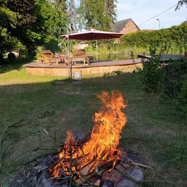 A lively campfire burns in the foreground, with a backyard deck, lounge chairs, sun umbrella, pond, and rustic building in a serene, green outdoor setting.