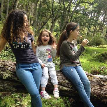 Das Bild zeigt drei junge Mädchen, die auf einem Baumstamm in einem Waldgebiet sitzen. Die beiden jüngeren Mädchen stehen vorne und das ältere Mädchen dahinter.