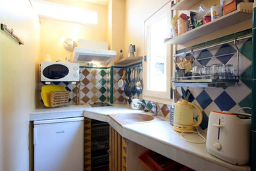 The image depicts a kitchen area with a sink, stove, and refrigerator, featuring a table and chairs in the background, with a window above the table. The room is well-lit and has a clean appearance.