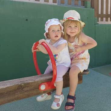 The image shows two young girls sitting on a wooden bench with a red toy, posing for the camera in front of a green wall.