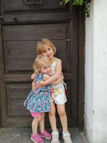 The image depicts two young girls standing in front of a wooden door, with one girl embracing the other from behind. They are both wearing dresses and sneakers.