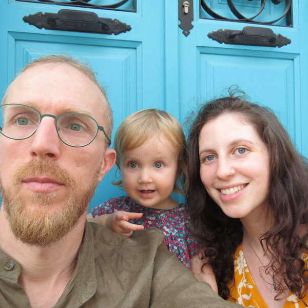The image depicts a family of three posing for a selfie in front of a bright blue door, with the man on the left, the child in the middle, and the woman on the right.