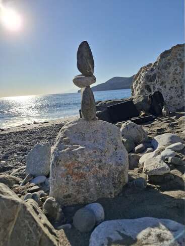 A serene beach scene shows a creative tower of balanced stones, sunlight glimmering on the ocean, rocky shore, backpack, and distant hills under a clear sky.