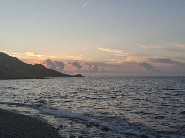 A serene beach scene at sunset, with gentle waves touching the shore, distant hills silhouetted, colorful clouds, and a faint trail from an airplane above. Peaceful atmosphere.