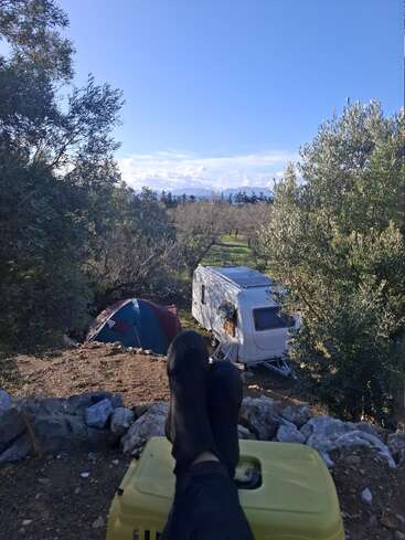A person relaxes with feet up, overlooking a peaceful campsite with a tent and caravan surrounded by olive trees under a bright blue sky.