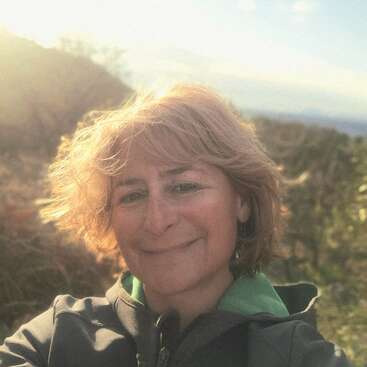 A woman with short blonde hair smiles outdoors. She wears a jacket and stands in sunlight, with a soft, dreamy background of nature and blue sky.